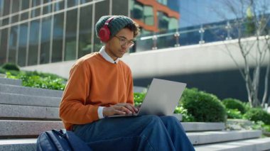 Kulaklık takan ve gözlüklü genç adam dışarıda merdivenlerde oturuyor, modern bir şehir ortamında dizüstü bilgisayarında daktilo ediyor..
