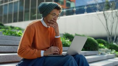 Genç adam dışarıda dizüstü bilgisayarla oturuyor, gözle görülür şekilde stresli ve derin düşünüyor, muhtemelen bir sorunla ya da zor bir seçimle karşı karşıya..