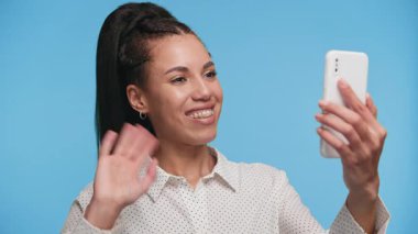 Gülümseyen genç bir kadın, mavi bir arkaplan karşısında akıllı bir telefon görüşmesinde sohbet ederken el sallıyor, bağlantı ve neşe gösteriyor..