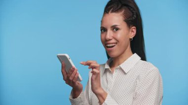 Heyecanlı kadın akıllı telefonu tutuyor ve alkışlıyor. Mavi arka planda tek başına, yumruğunu kaldırmış.