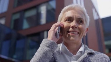 Modern bir ofis binasının önünde akıllı telefondan konuşan mutlu yaşlı kadın.
