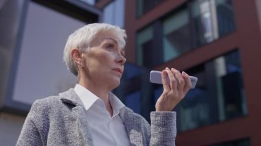 Modern bir ofisin dışında sesli mesaj kaydetmek için telefona konuşan odaklanmış yaşlı bir kadın.