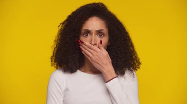 Şaşırmış kadın, geniş gözlü, ağzı kapalı, elleri parlak, sarı arka planda.