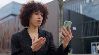 İnce çizgili takım elbiseli mutlu genç bir kadın modern ofis binasının dışındaki akıllı telefonuyla yaptığı bir video görüşmesi sırasında gülümsüyor..