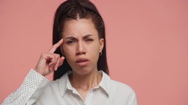 Genç bir kadın kafasını şüpheli bir ifadeyle işaret ederek güvensizlik işareti mi veriyor yoksa pembe arka planda duran benekli bir tişörtle çılgınca bir jest mi yapıyorsun?.