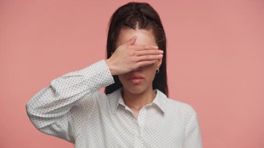 Puantiyeli tişörtlü, ciddi bir genç kadın tek eliyle gözlerini kapatıyor. İnanmadığını, hayal kırıklığına uğradığını ya da pembe arka plandan kaçındığını ifade ediyor..