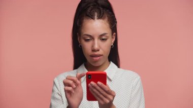 Puantiyeli tişörtlü neşeli genç bir kadın pembe arka planda akıllı telefonundan iyi haberler okurken ışıl ışıl gülümser..