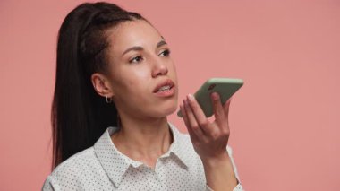Puantiyeli tişörtlü odaklanmış bir kadın akıllı telefonunu yakın tutuyor ve pembe arka planda bir sesli mesaj kaydediyor..