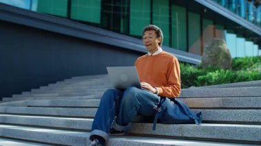 Turuncu kazaklı ve bereli genç bir üniversite öğrencisi, dışarıda kampüs merdivenlerinde otururken laptopuyla bir video görüşmesine katılıyor..