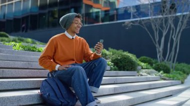 Turuncu kazaklı ve bereli neşeli öğrenci kampüs merdivenlerinde oturmuş telefonunda neşeli bir video görüşmesi yapıyor, gülümsüyor ve nişanlanıyor..