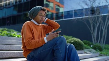 Şaşırmış ve endişeli bir öğrenci açık hava basamaklarında oturuyor, şok ve endişe içinde, akıllı telefondan beklenmedik ya da kötü haberler okurken..