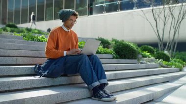 Odaklanmış genç adam elinde dizüstü bilgisayarla kentsel basamaklarda oturuyor, yeşillik ve binalarla çevrili modern bir açık havada uzaktan çalışıyor..