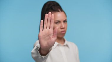 Kendine güvenen genç bir kadın kameranın önünde el kaldırıyor, dur işareti veriyor ya da reddediyor mavi arka planda sert bir yüz ifadesiyle..