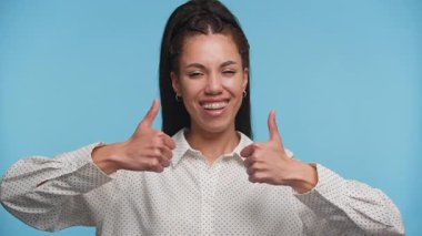 Kendine güvenen, gülümseyen, iki parmağını havaya kaldıran, onay veren, pozitiflik ve mavi arka plana karşı cesaretlendiren genç bir kadın..