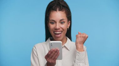 Çok neşeli genç bir kadın akıllı telefona bakıyor ve bir yumruk pompasıyla tezahürat yapıyor, mavi arka plandaki heyecan verici ya da olumlu haberlere tepki veriyor..
