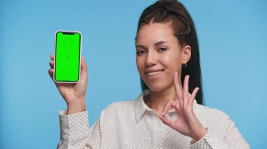 Mutlu genç bir kadın yeşil ekranlı akıllı telefonu sunuyor ve iyi bir jest yapıyor. Uygulama promosyon kavramları için ideal, mavi arka planda izole edilmiş..