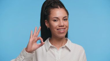 Gülümseyen genç bayan, güvenle işaret dilini gösteriyor. Onayını, pozitifliğini ve mavi arka plandan memnuniyetini ifade ediyor..