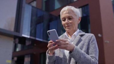 Profesyonel kıyafetli, kendinden emin yaşlı bir kadın modern bir ofis binasının dışındaki akıllı telefonuna bir mesaj yazar..