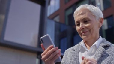 Açık havada akıllı telefon kullanarak gülümseyen olgun bir kadın, muhtemelen modern bir şehir ofis binasının önünde internette geziniyor ya da alışveriş yapıyor..