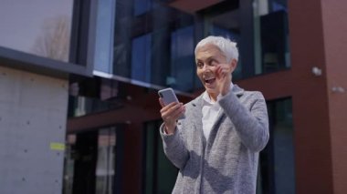 Dışarıdaki neşeli yaşlı kadın akıllı telefonuna bakıp gülerken bir mesaja olumlu tepki veriyor..