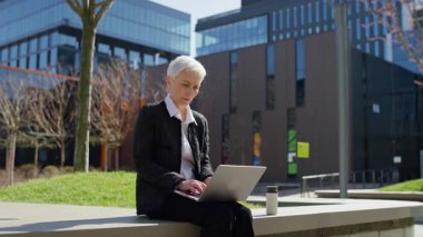 Odaklanmış yaşlı kadın dışarıda dizüstü bilgisayarla oturuyor, arka planda ofis binaları olan modern bir şehir ortamında çalışıyor..