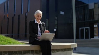 Mutlu yaşlı bir kadın bilgisayarında video görüşmesi yaparken el sallıyor güneşli gökyüzünün altındaki bir ofis parkında çalışıyor..
