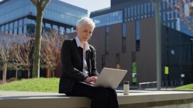 Düşünceli olgun bir kadın dışarıda dizüstü bilgisayarla oturur ve modern ofis alanındaki önemli bir kararı düşünmek için işi durdurur..