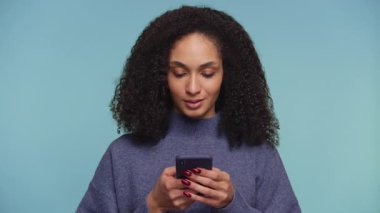 Odaklanmış kadın telefona düşünceli bir ifadeyle bakar, mesaj atarken cevap vermeyi düşünür, düz mavi bir arkaplana karşı..