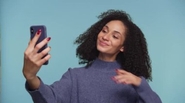 Kendine güveni tam genç bir kadın bir selfie için poz veriyor, tek eliyle öpücüklü bir yüz ve barış işareti yapıyor, sağlam bir mavi arka plana karşı..