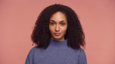 Sakin ve kendine güvenen mavi kazaklı bir kadının portresi. Yumuşak pembe bir arka planda nötr bir bakışla duruyor..