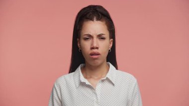 Üzgün ya da üzgün görünen genç bir kadının portresi stüdyo ışıklarında yumuşak pembe bir arka plana karşı dururken hafifçe kaşlarını çatıyor..
