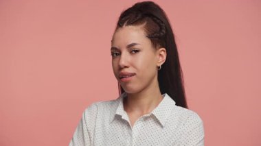 Kendine güveni tam genç bir kadın kameraya şakacı bir şekilde göz kırpıyor, yumuşak pembe bir stüdyonun arka planında duruyor ve kurnazca gülümsüyor..