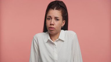 Genç kadın şaşkın ve hafifçe omuz silkiyor, belirsizlik ve şaşkınlık ifade ediyor, stüdyoda düz pembe bir arka plan karşı.