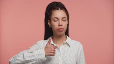 Tarafsız bir yüz ifadesi takınırken, pembe bir zemine karşı duran genç bir kadın başparmak hareketi yapıyor..