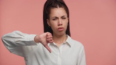 Onaylamayan bir ifadeyle baş parmak gösteren genç bir kadın stüdyoda pembe bir arka plana karşı duruyor..