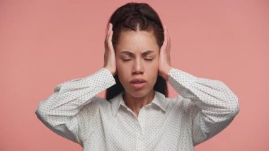 Stres ya da hayal kırıklığı gösteren genç bir kadın gözleri kapalı ve elleri kulaklarının üzerinde stüdyoda pembe bir arka plana karşı duruyor..
