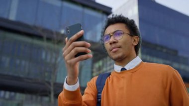 Turuncu kazaklı ve gözlüklü bir genç modern şehir ortamında akıllı telefonunu kullanarak bir video görüşmesinde konuşuyor..
