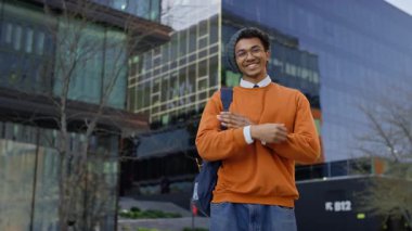 Modern bir kampüs binasının önünde gülümseyerek poz veren kendine güvenen genç bir öğrenci, gündelik kıyafetler ve sırt çantası giyiyor..