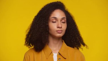 Kıvırcık saçlı genç bir kadının portresi. Parlak sarı bir arka planın önünde güvenle duruyor. Doğrudan kameraya bakıyor..