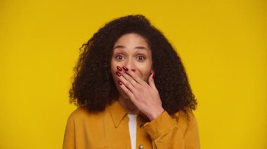 Geniş gözlü genç bir kadın şok ya da şaşkınlıkla ağzını kapatıyor. Stüdyoda canlı sarı bir arka plana karşı duruyor..