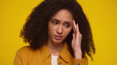 Genç bir kadın endişeli görünüyor ve şakağına dokunuyor. Parlak sarı bir arka planda stres ya da endişe belirtileri gösteriyor..