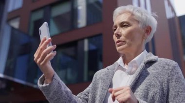 Kendinden emin yaşlı bir kadın, iletişim ve bağlantıyı temsil eden modern iş binasının dışındaki bir video görüşmesi için akıllı telefonu kullanıyor..