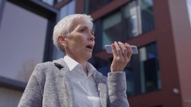 Olgun bir iş kadını, modern bir ofis binasının dışında dijital iletişimi gösteren bir sesli mesaj kaydetmek için akıllı telefona konuşuyor..