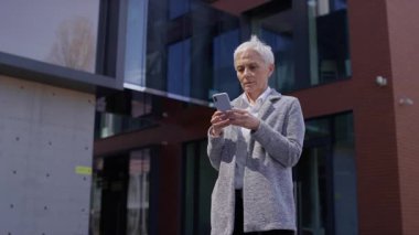 Modern bir ofis binasının önünde akıllı telefonuna mesaj yazarken gülümseyen yaşlı, profesyonel bir kadın dijital bağlantıyı yansıtıyor..