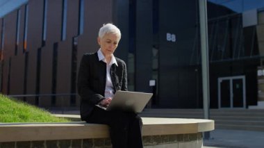 Modern bir iş bölgesinde olumlu haberler okurken dışarıda dizüstü bilgisayarla oturan mutlu, yaşlı ve profesyonel bir kadın..