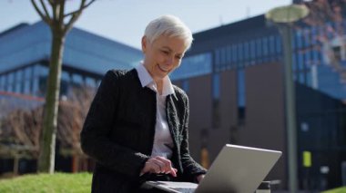 Yaşlı profesyonel bir kadın modern bir şehir ortamında oturuyor, iş molasında laptopunu karıştırırken gülümsüyor..