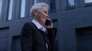 Neşeli, yaşlı, profesyonel bir kadın karanlık cam binalarla modern bir iş bölgesinde yürürken keyifli bir telefon görüşmesi yapıyor..