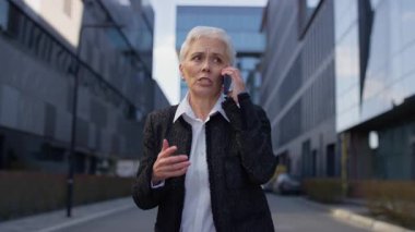 Endişeli bir ifadeyle yaşlı bir iş kadını modern bir şehir bölgesinde kulağına bir telefon tutarak stres veya kötü haberi işaret ediyor..
