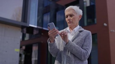 Yaşlı profesyonel bir kadın akıllı telefonunu yüzünde tutarak üzücü bir haber ya da mesaj gördükten sonra kaygılanarak tepki veriyor..
