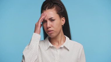 Hüsrana uğramış bir ifadeye sahip genç bir kadın. Alnını stres ya da şaşkınlık içinde tutuyor. Duygusal kavramlar için mavi bir arkaplan..